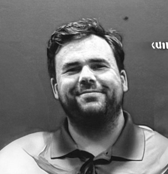 Close-up portrait of a young man with dark hair and stubble, smiling, wearing a collared shirt