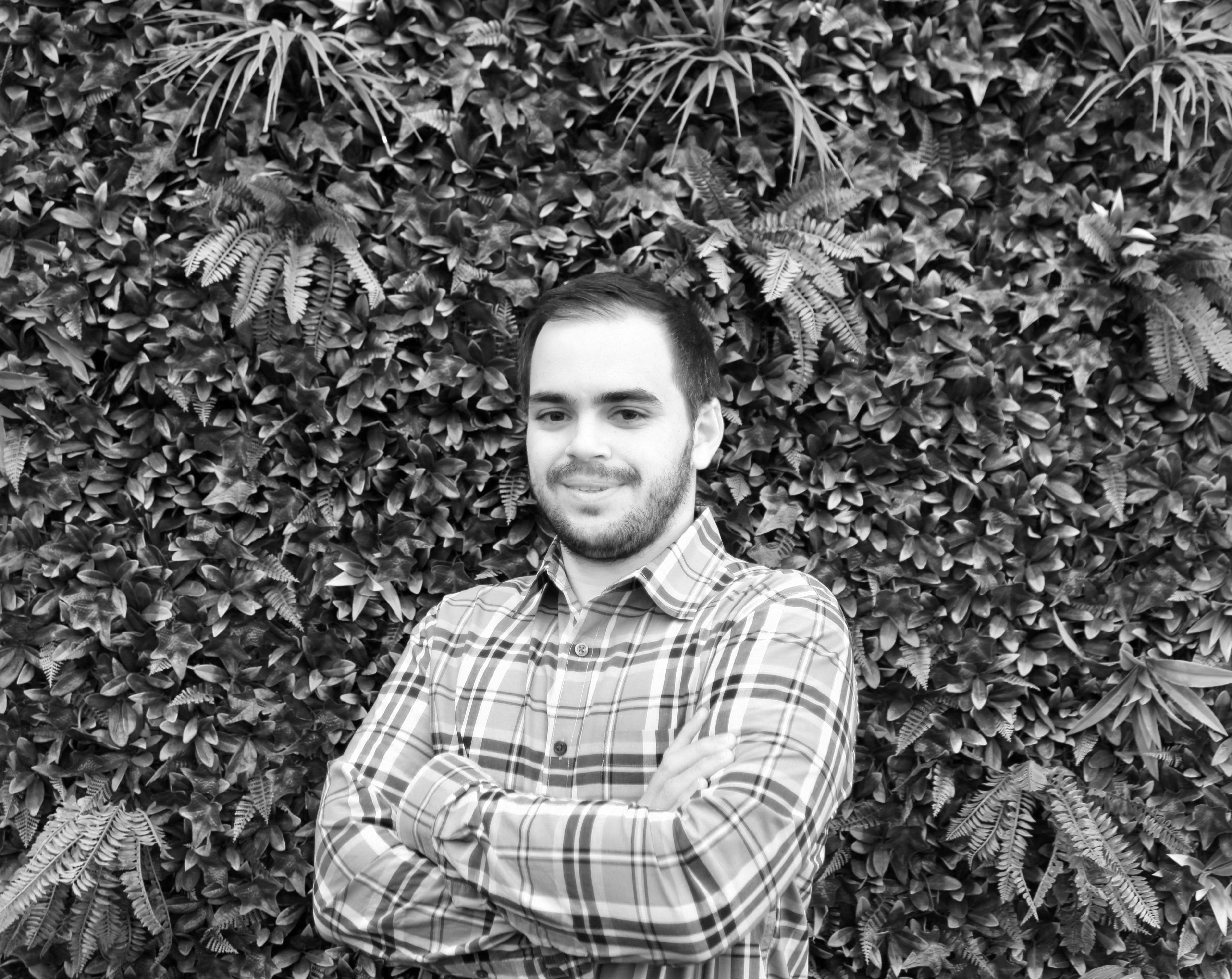 Portrait of a young man standing against a wall of dense foliage, wearing a flannel shirt.