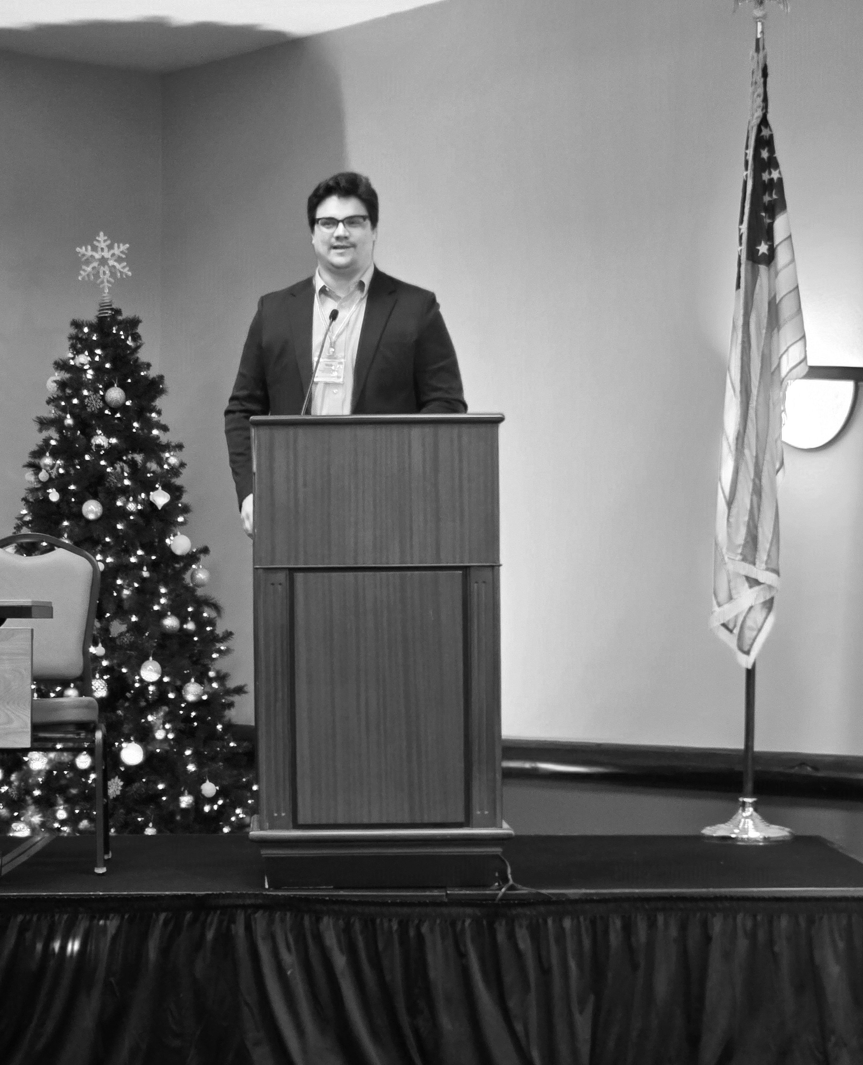 Portrait of Nick, delivering a speech behind a podium at a conference.
